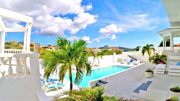 Un área de piscina con sillones de descanso y una palmera, rodeada de edificios blancos y un cielo despejado.