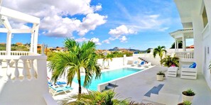 Un área de piscina con sillones de descanso y una palmera, rodeada de edificios blancos y un cielo despejado.