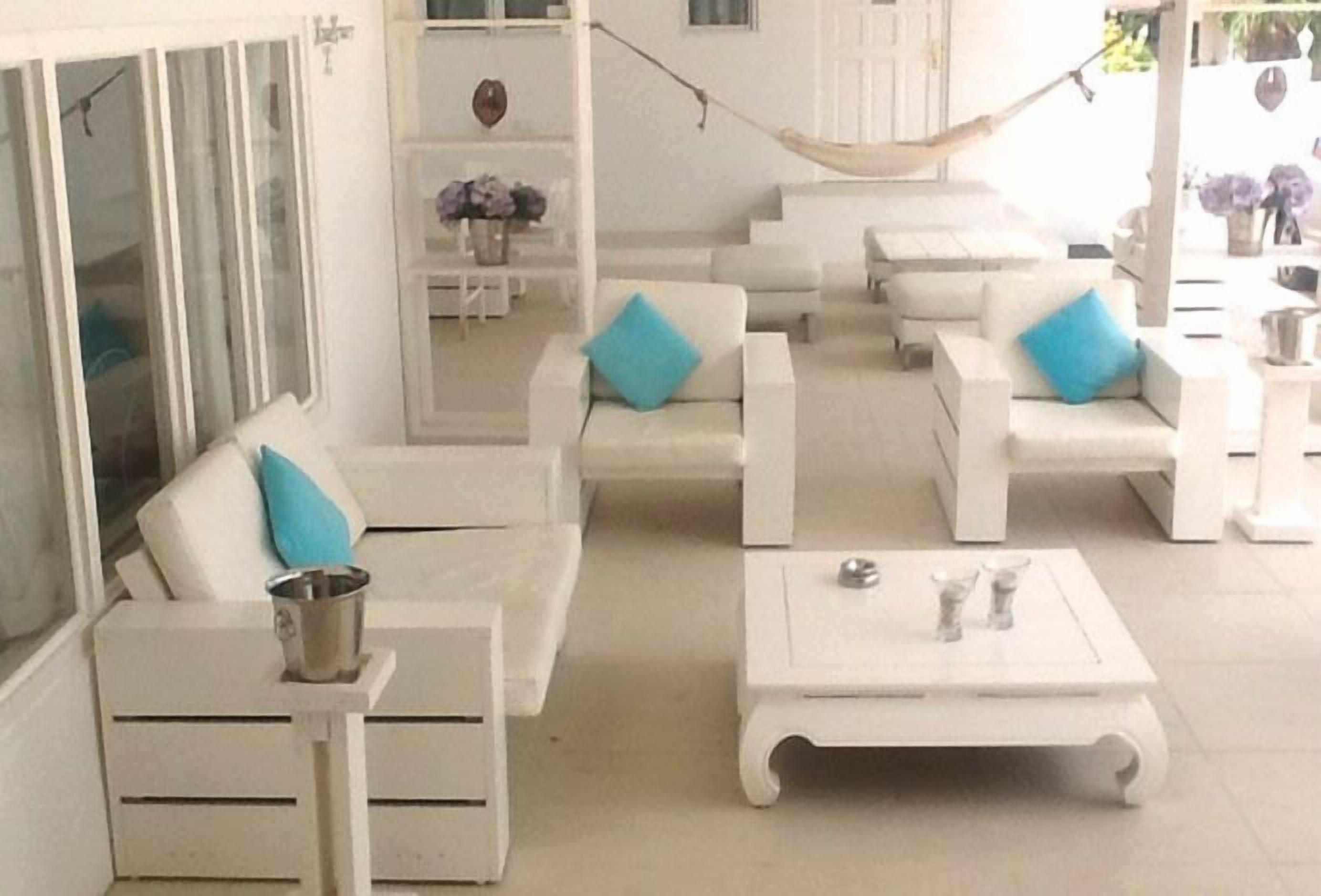 Un área de estar exterior blanca con sillas acolchadas y una mesa, una hamaca y un armario blanco con un jarrón de flores.