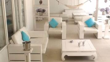 Un área de estar exterior blanca con sillas acolchadas y una mesa, una hamaca y un armario blanco con un jarrón de flores.