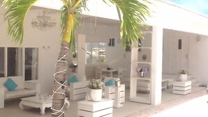 Un área de descanso al aire libre moderna con muebles blancos, una palmera y un edificio blanco con puerta de vidrio.