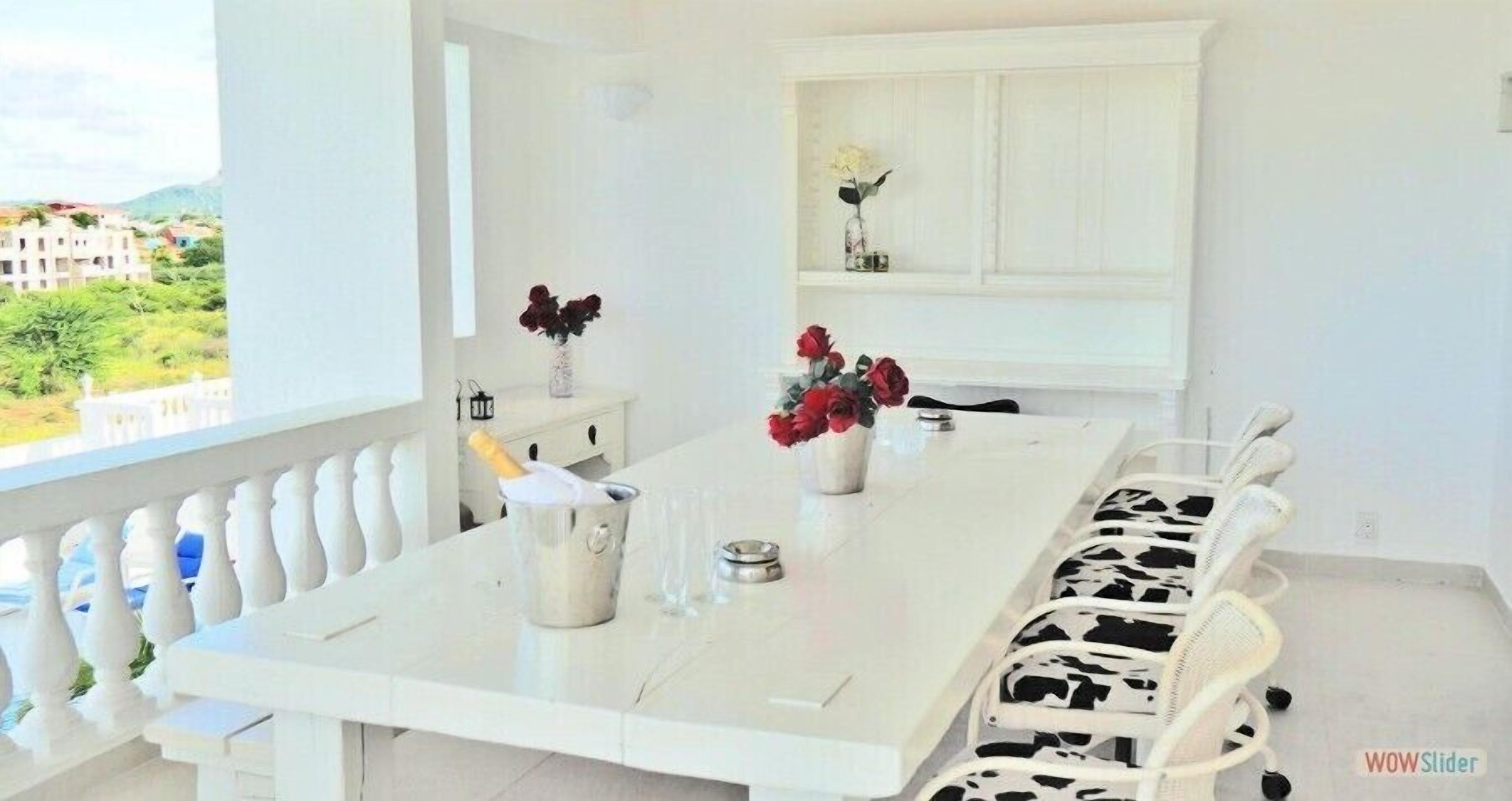 Un comedor moderno con una mesa y sillas blancas, un jarrón con flores rojas y vistas a edificios.