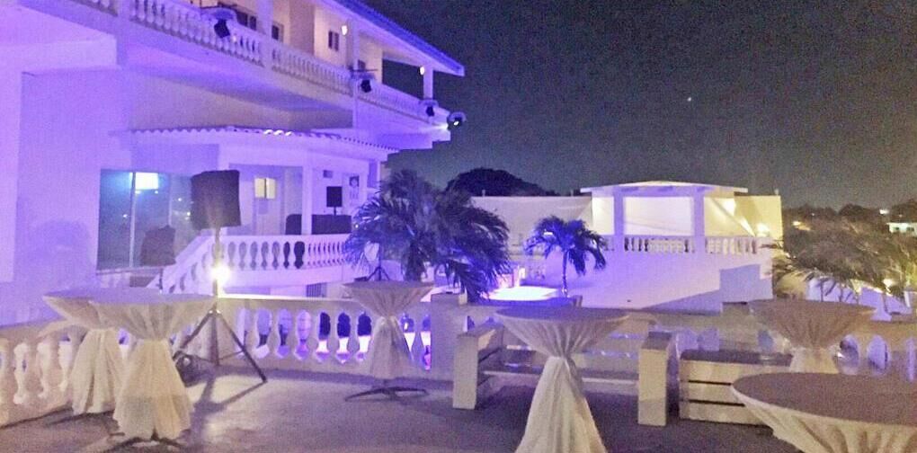 Un espacio al aire libre con mesas y sillas, un edificio con balcones y palmeras.