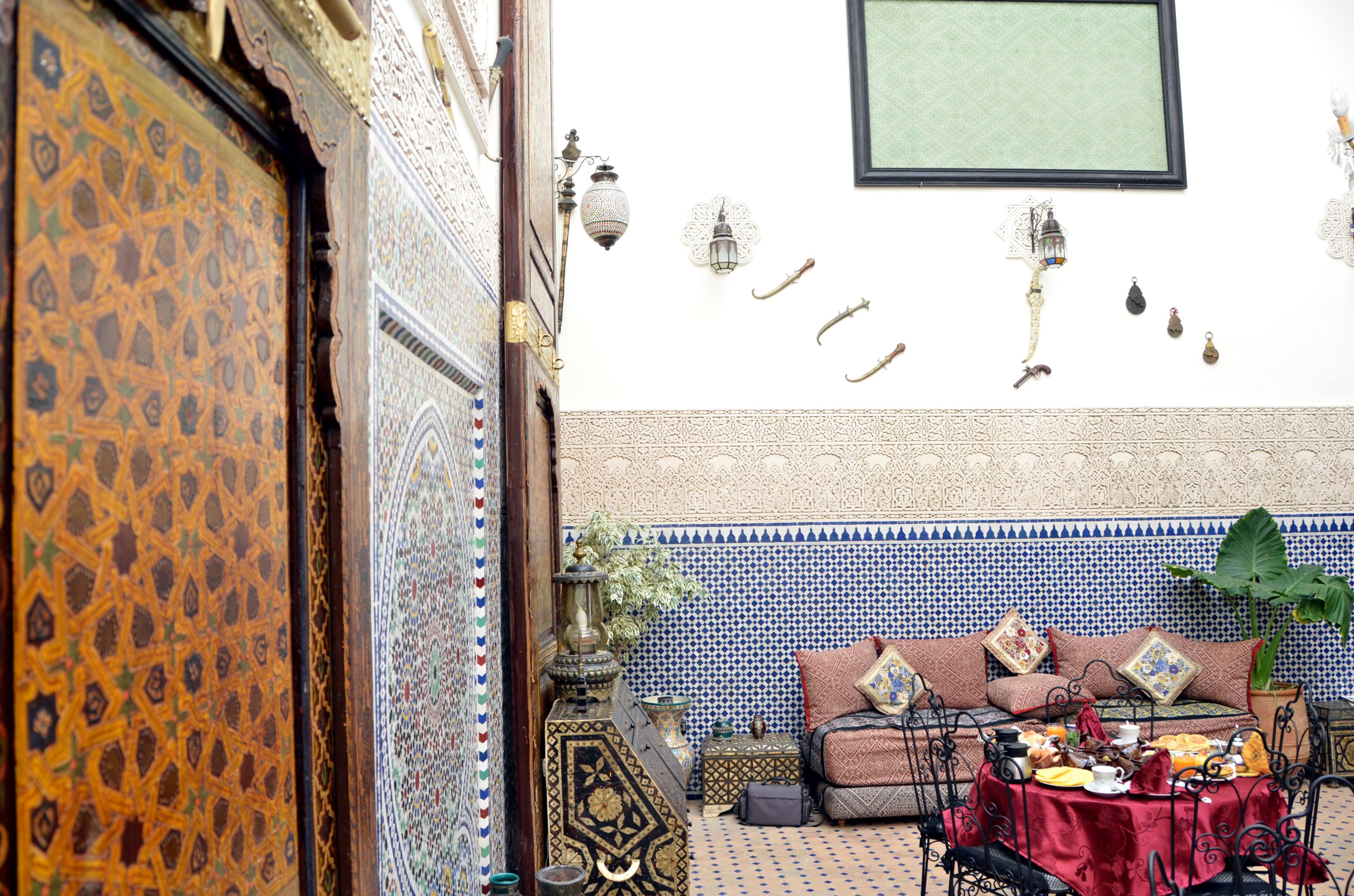 A traditional Moroccan interior with a patterned door, a tiled wall, a sofa with cushions, and a table set for a meal.