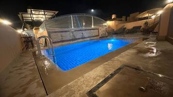 A rooftop swimming pool with a glass enclosure, lounge chairs, and a covered seating area.
