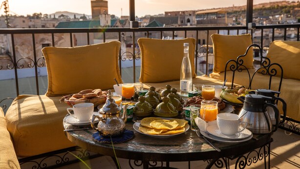 A balcony with yellow cushions, a table set with breakfast items, and a view of a cityscape in the background. - Riad Dar Tahri & SPA (Fes)