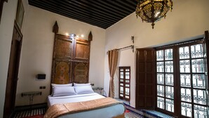 A traditional Moroccan room with a bed, patterned floor, ornate wooden cabinet, and a large window with iron bars. - Riad Dar Tahri & SPA (Fes)