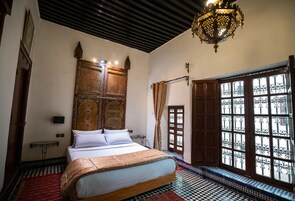 Quarto marroquino tradicional com cama, piso estampado, armário de madeira ornamentado e uma janela grande com grades de ferro.