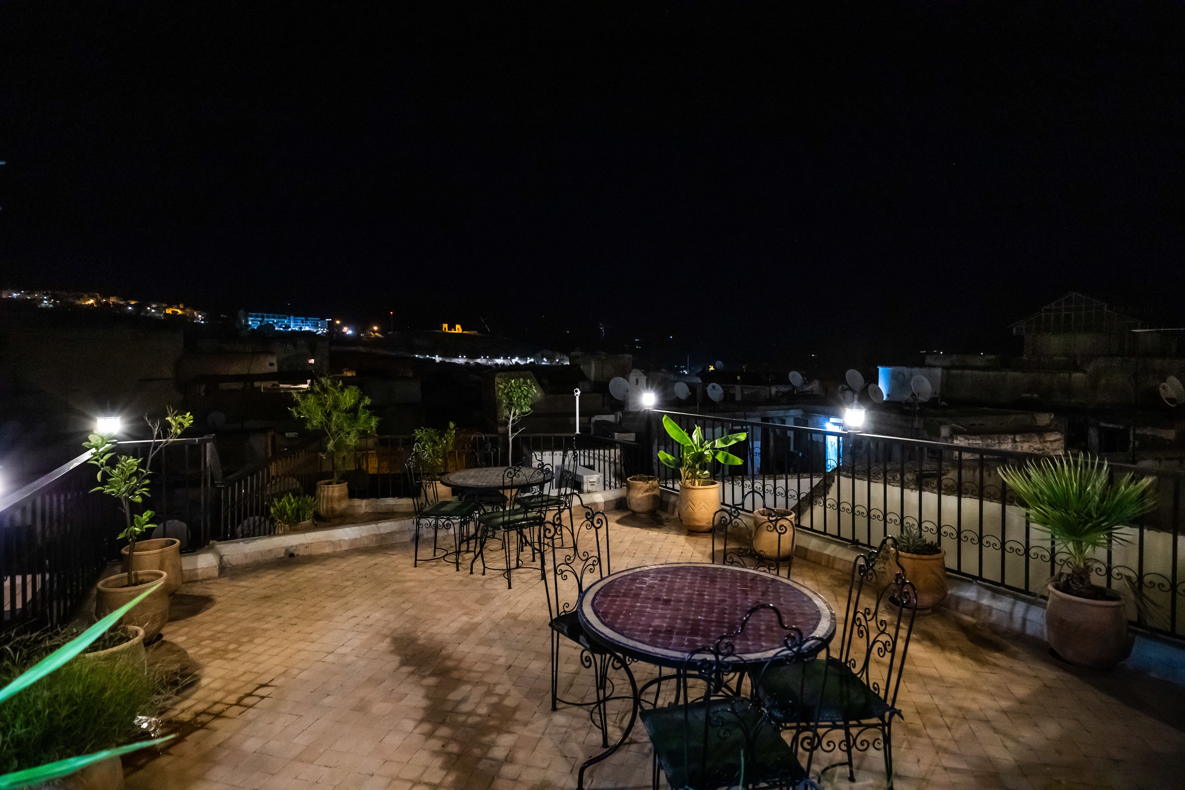 Un patio al aire libre con piso de ladrillo, mesas y sillas de metal, plantas en macetas y una barandilla.
