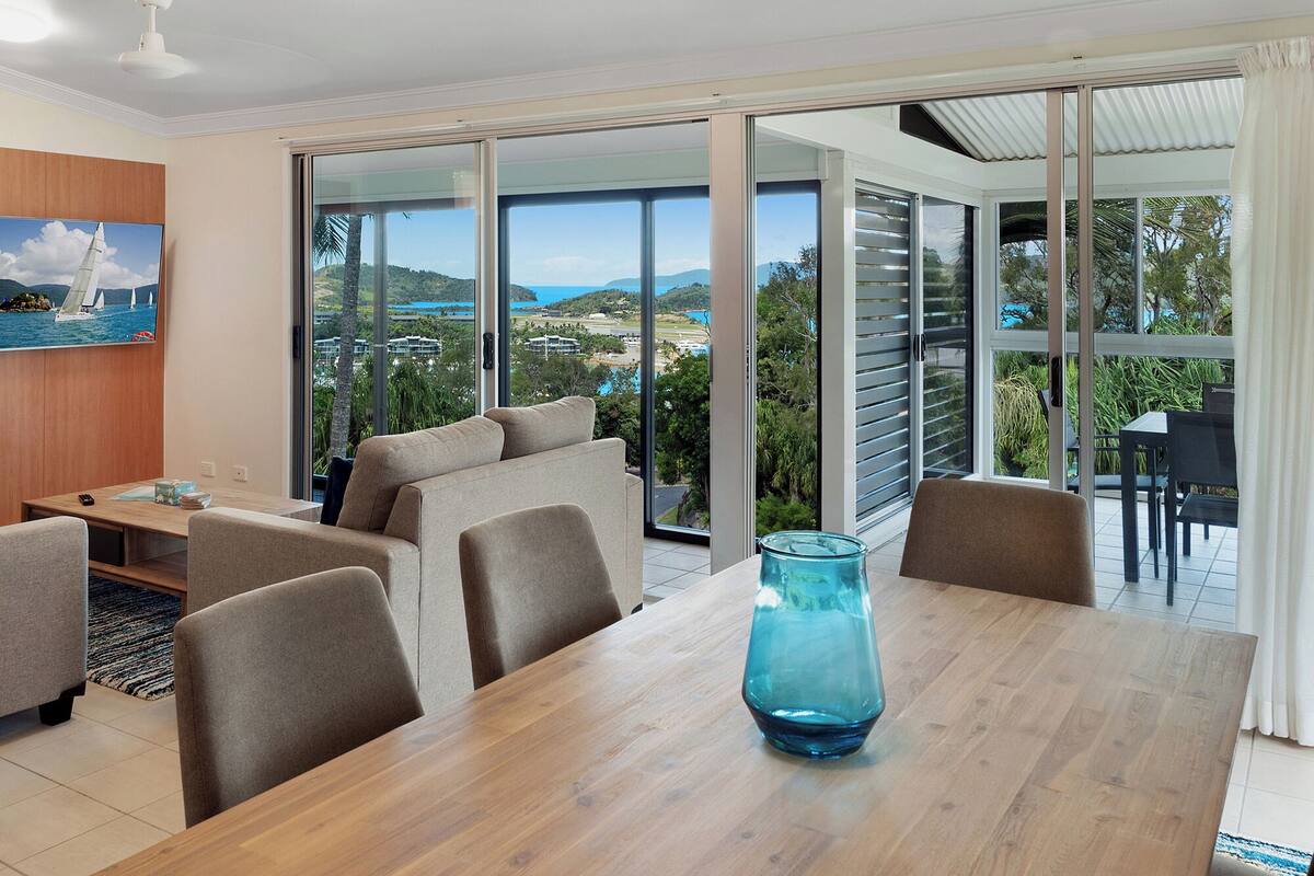 A modern room with a large sliding glass door leading to a balcony with a view of the sea and hills.