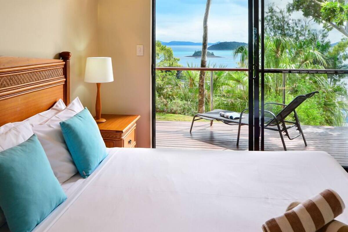 A hotel room with a bed, pillows, a nightstand with a lamp, and a view of a balcony with a chair.