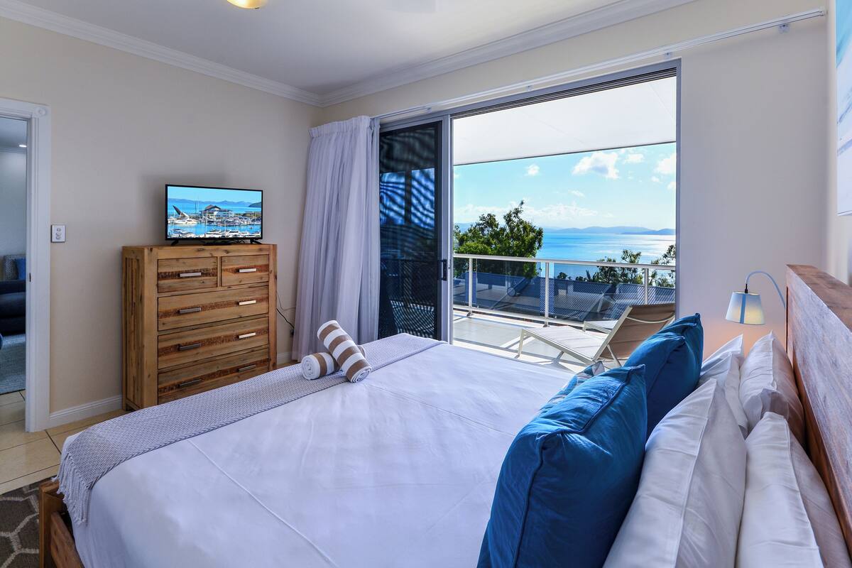 A bedroom with a bed, a wooden dresser, a TV, and a balcony with a view of the sea.