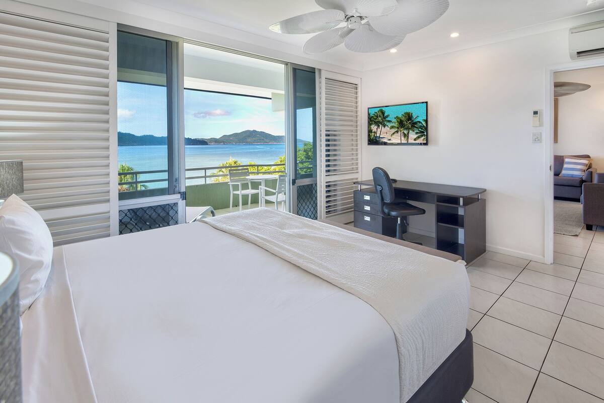 A bedroom with a bed, a desk, and a view of the sea.
