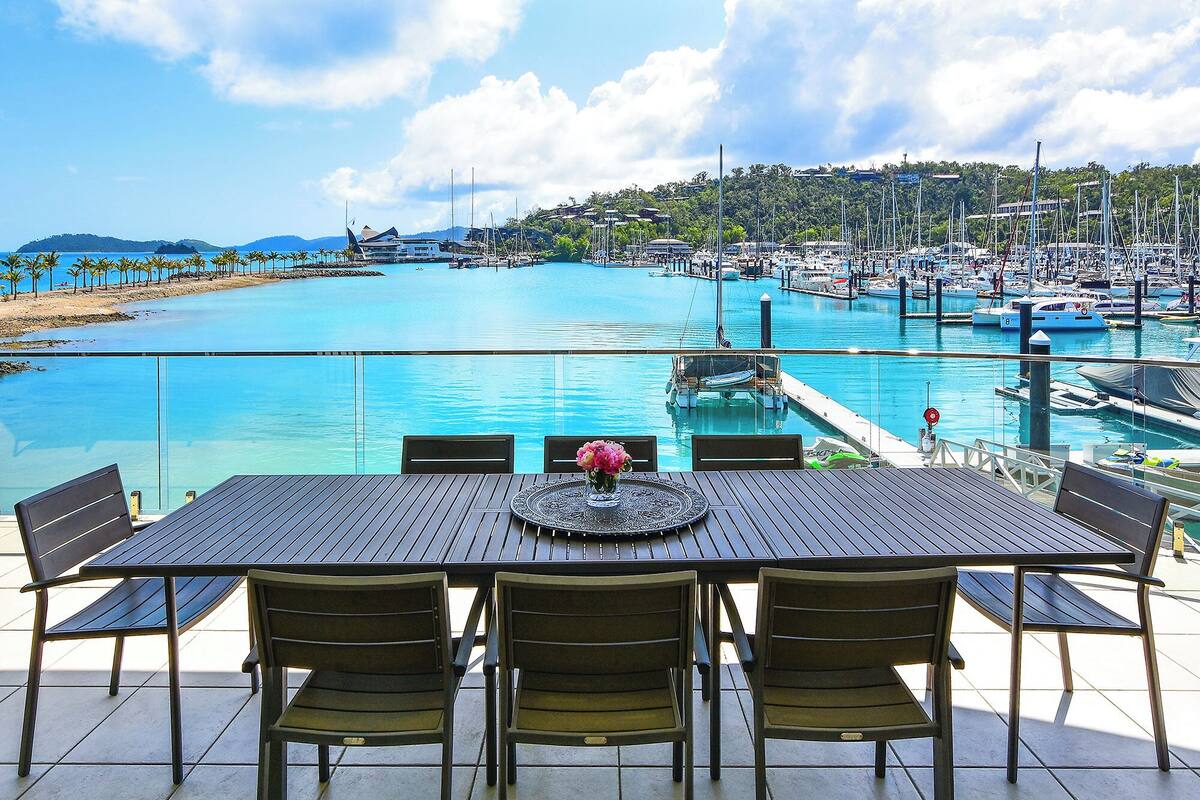 A dining table with chairs and a vase of flowers, overlooking a marina with boats and a beach.