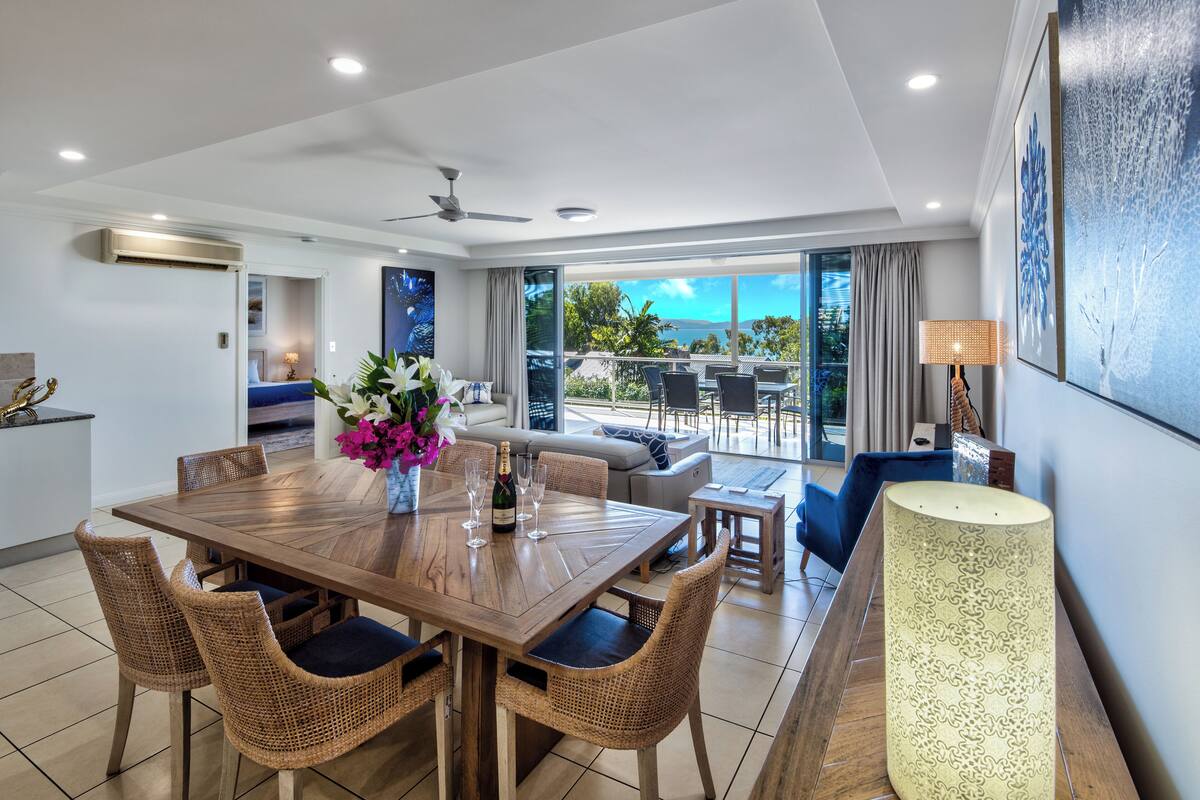 A spacious dining area with a wooden table and wicker chairs, a sofa, and a large window offering a view of the outdoors.