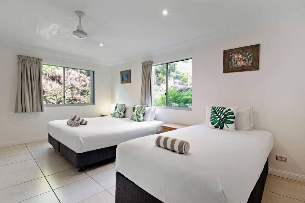 A hotel room with two beds, a ceiling fan, large windows, and a painting on the wall.