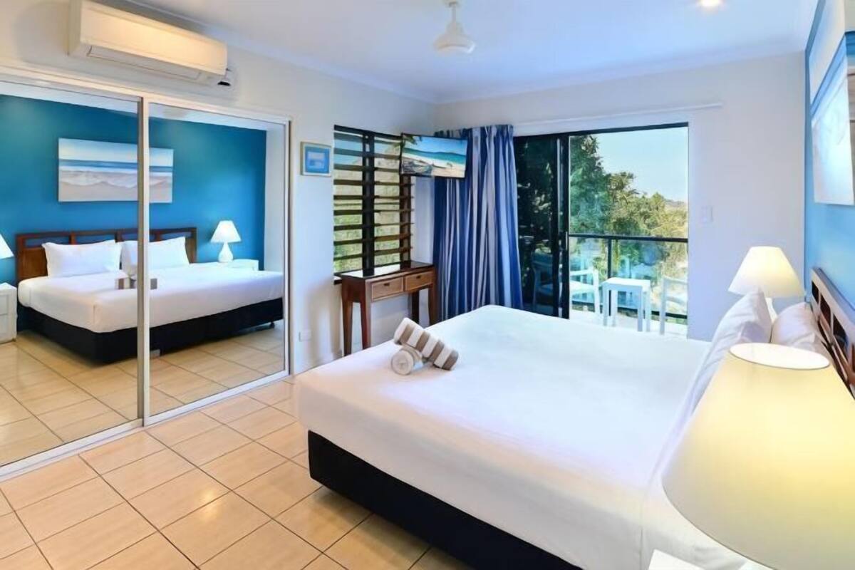 A hotel room with a large bed, a desk, a chair, a balcony with a table and chairs, and a sliding glass door.