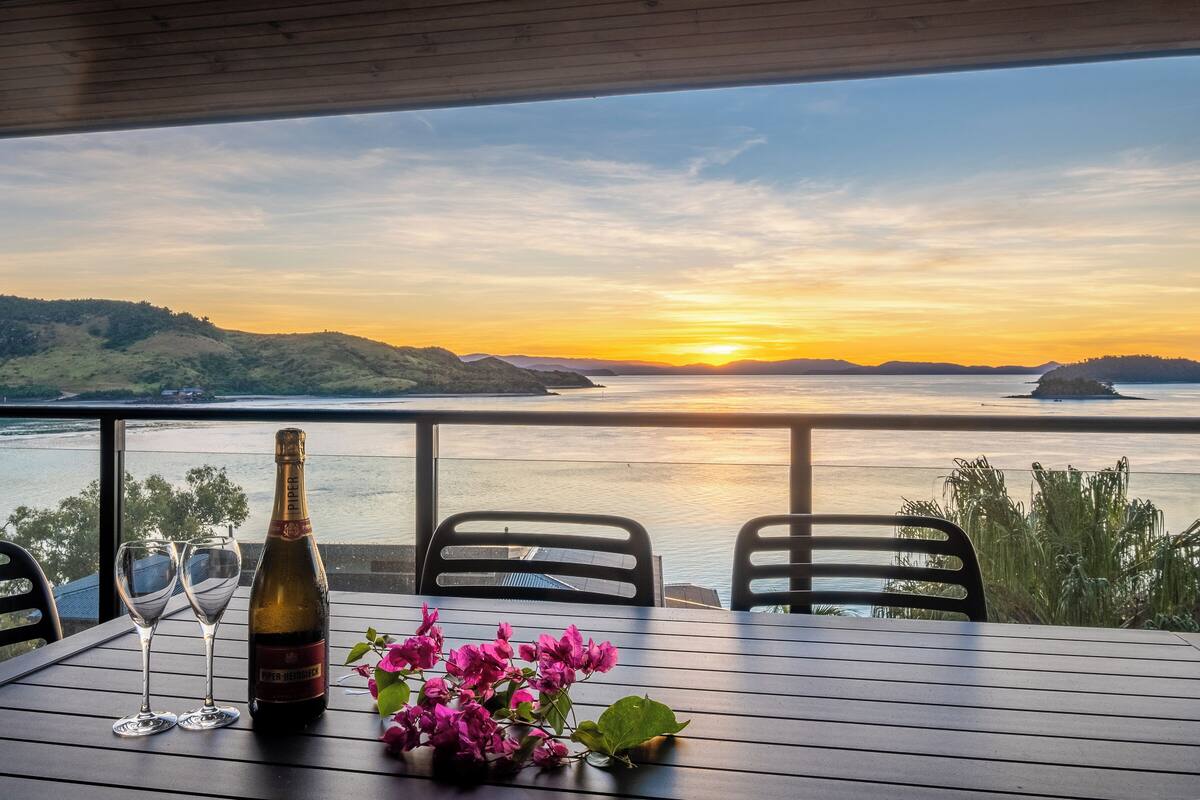 A table with a bottle and glasses, a vase of flowers, and a view of a sunset over water.