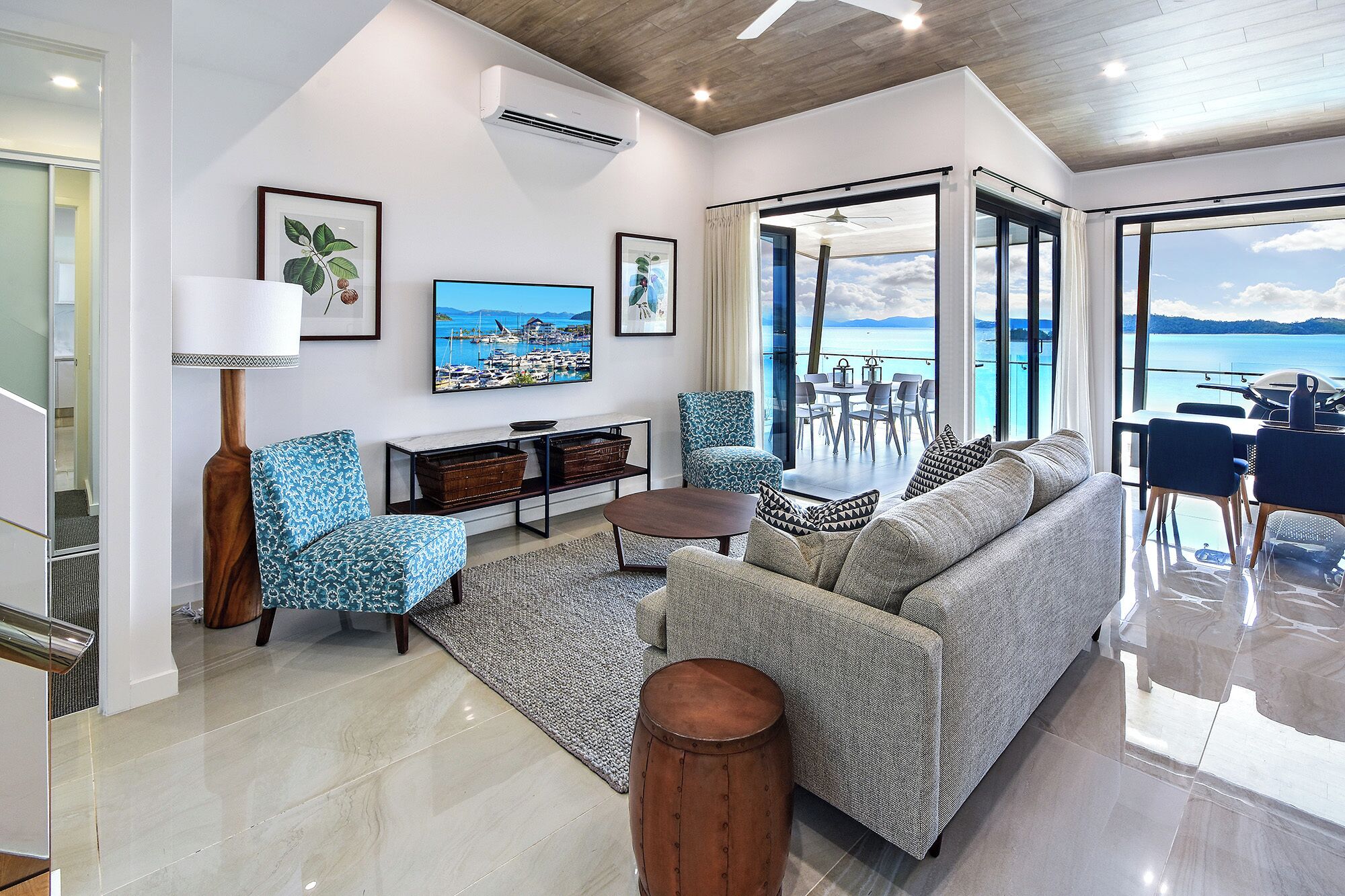A modern living room with a sofa, a coffee table, a patterned armchair, a side table, and a wooden stool. There are framed pictures on the wall, a ceiling fan, and a view of the sea through large windows.
