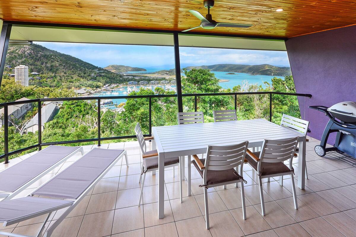 A balcony with a table and chairs, a grill, and a view of a coastal landscape.
