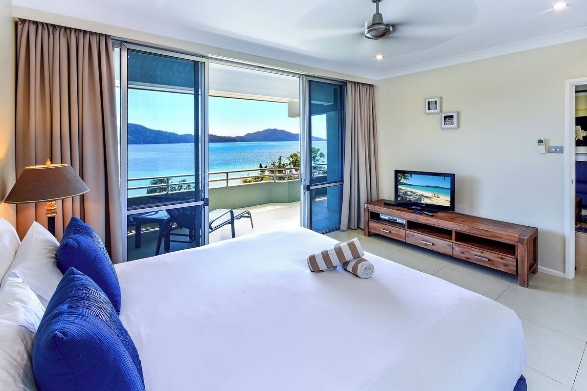 A hotel room with a large bed, a TV, a balcony with a view of the sea, and a sliding door.
