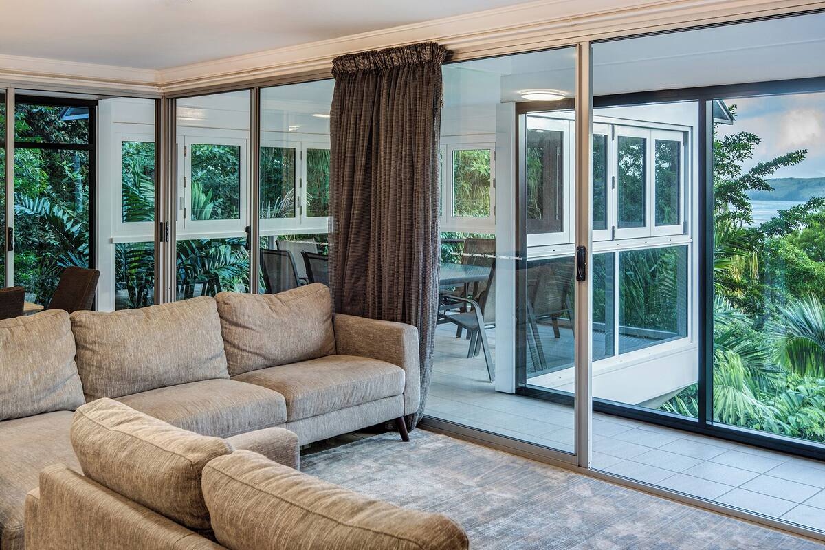 A modern living room with a large sofa, a dining area with chairs, and a view of lush greenery through large glass doors.