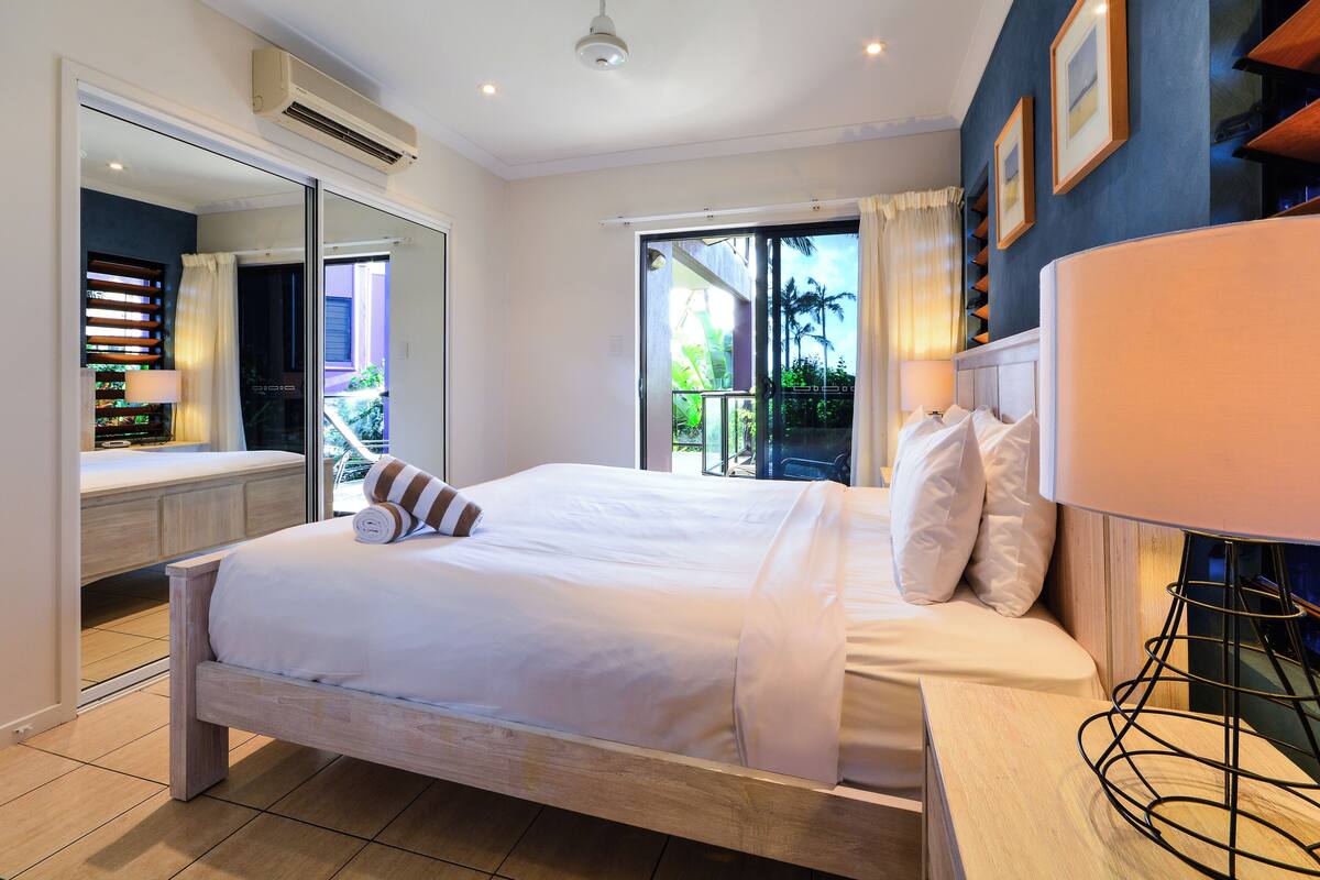 A bedroom with a bed, a bedside table, a mirror, and a view of the outdoors.