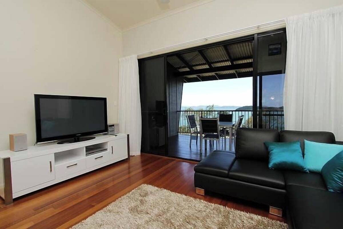 A modern living room with a flat-screen TV on a white console, a black leather sofa with teal cushions, and a large sliding glass door leading to a balcony with a view of the mountains.