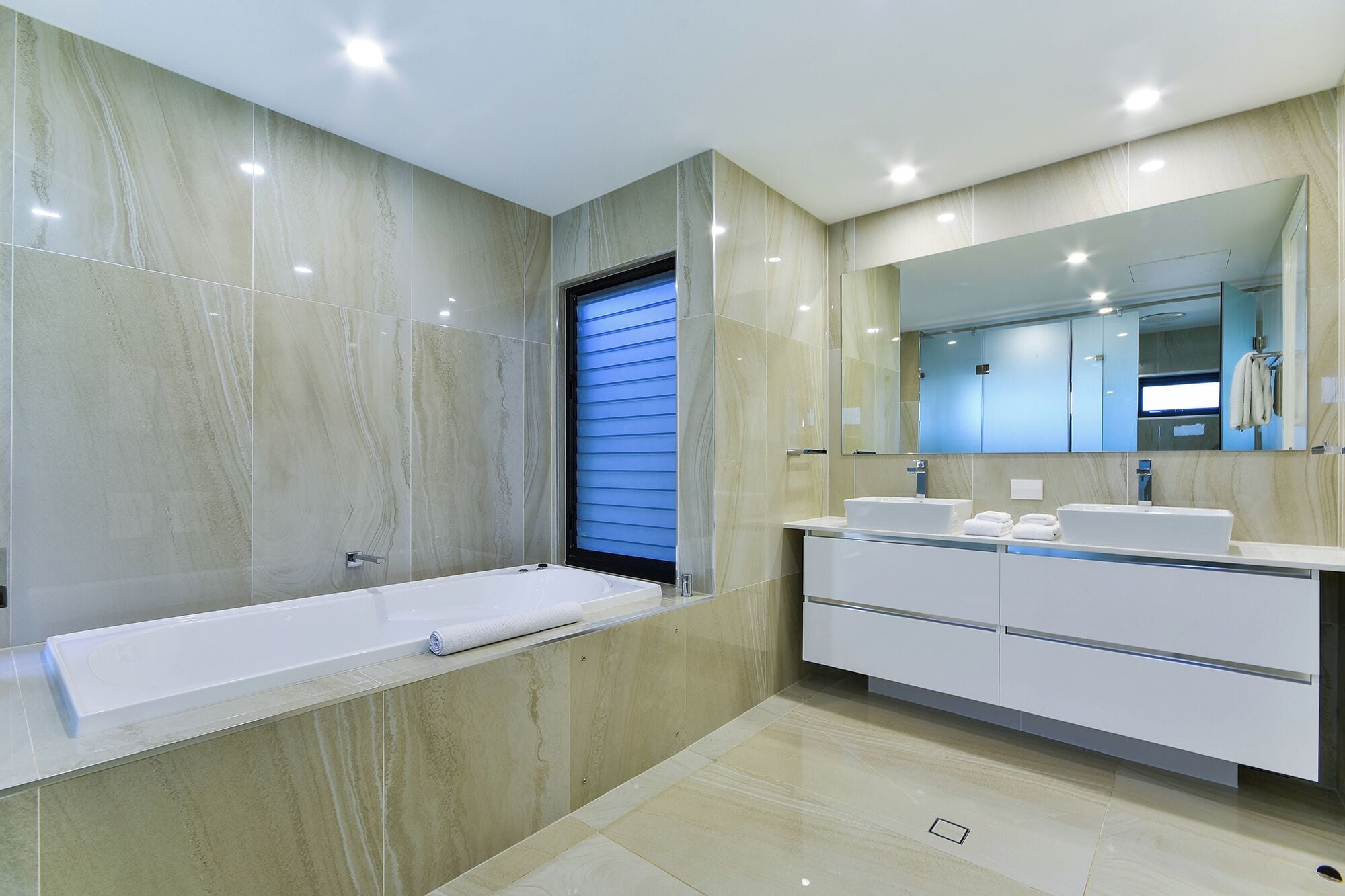 A modern bathroom with a bathtub, two sinks, a large mirror, and a window with blinds.