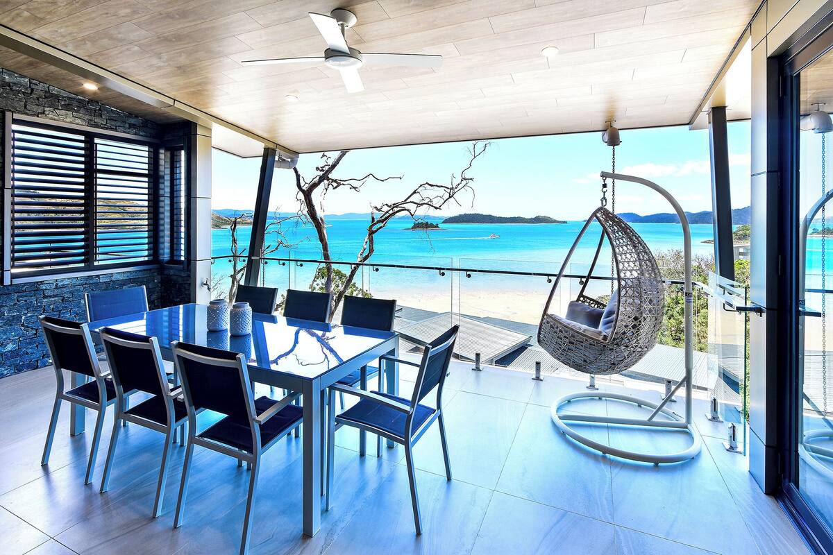A modern outdoor dining area with a view of the sea, a hanging chair, and a ceiling fan.