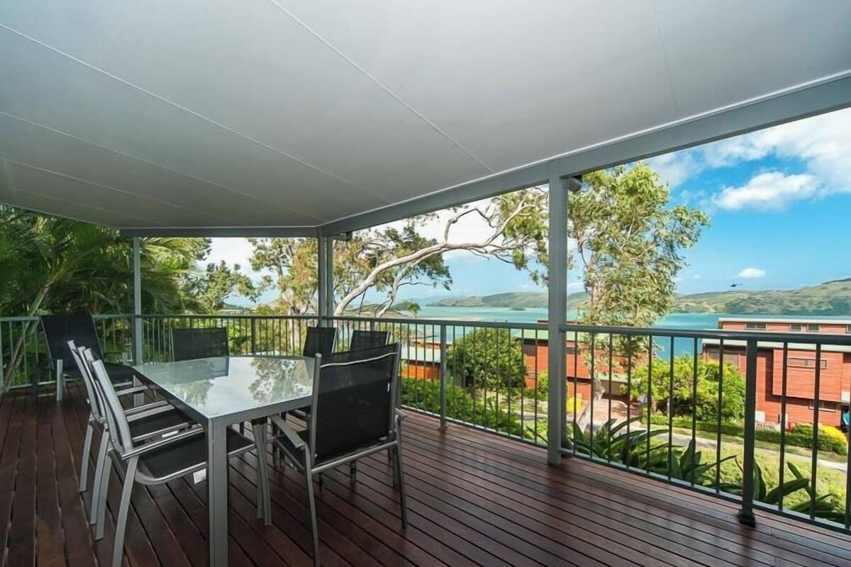 A covered outdoor area with a table and chairs, a view of a coastal landscape, and a metal railing.