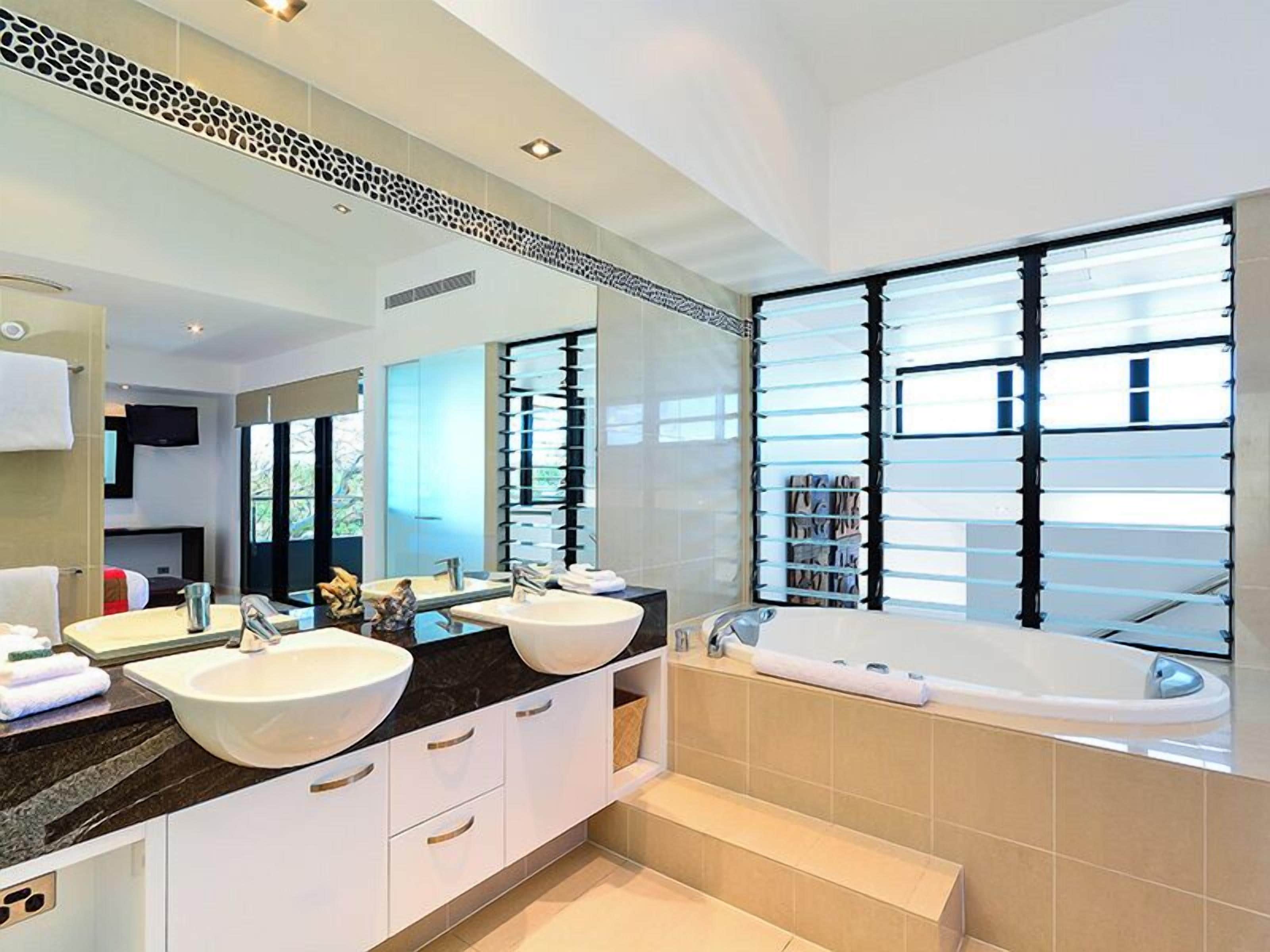 A modern bathroom with two sinks, a large mirror, a bathtub, and a view of the outdoors.