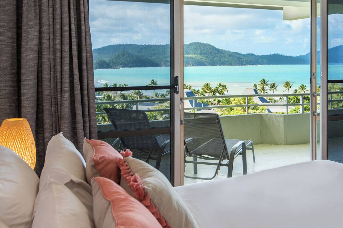A hotel room with a bed, a view of the beach, and a balcony with chairs.