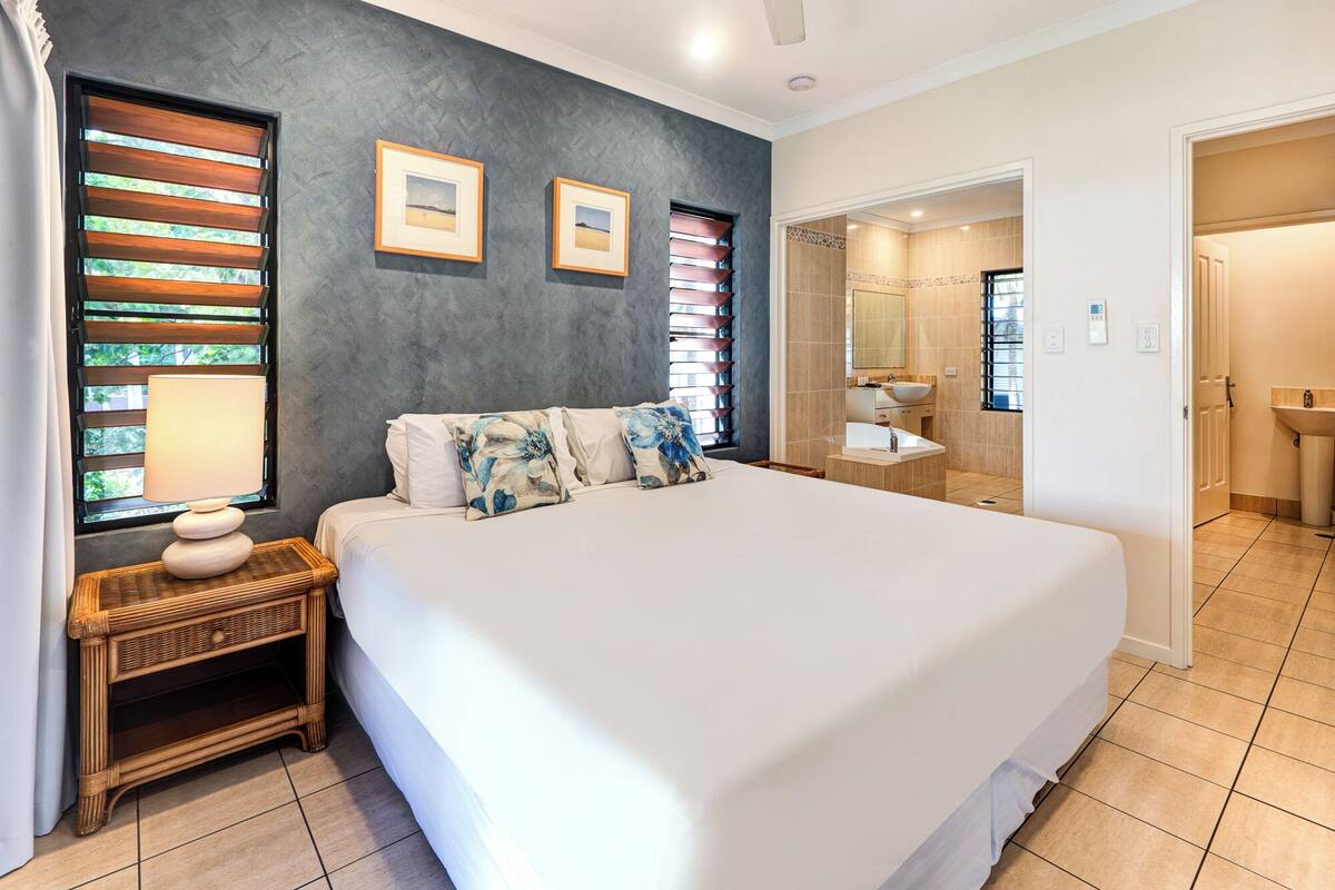 A bedroom with a large bed, a bedside table with a lamp, a wall-mounted picture, and a bathroom with a sink and mirror.