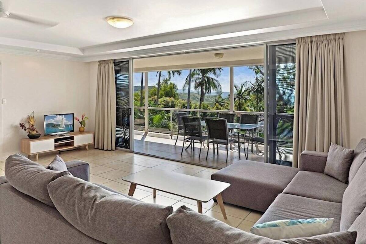 A modern living room with a sofa, a coffee table, and a TV on a wooden stand. There is a balcony with a table and chairs, and a view of palm trees and mountains.