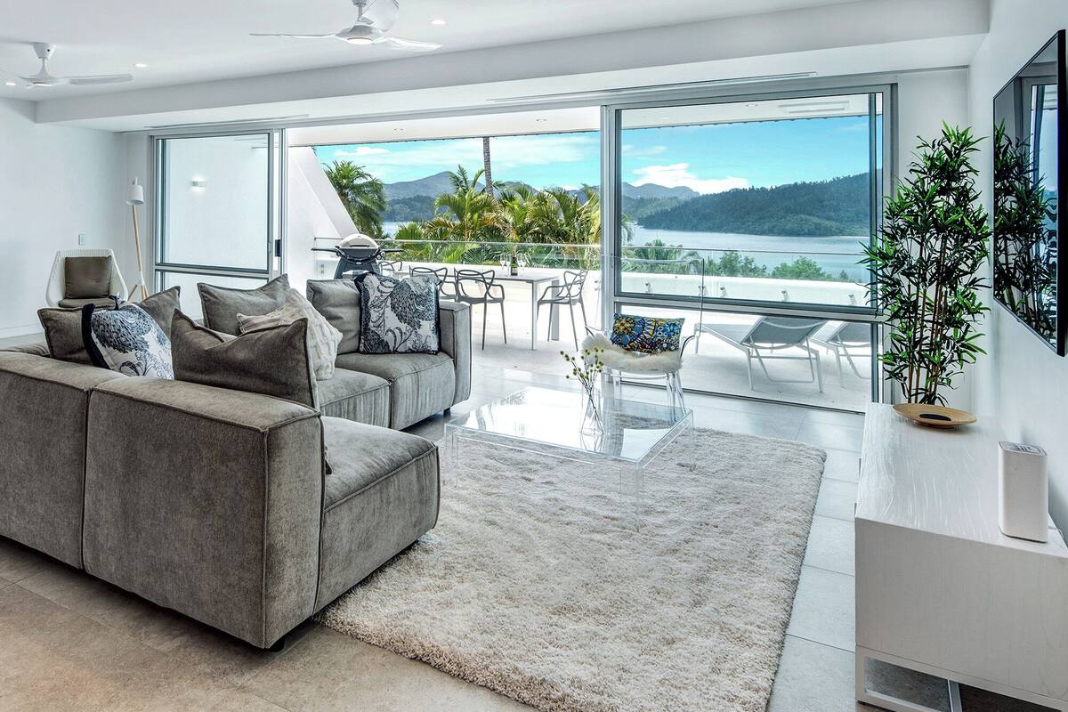 A modern living room with a grey sofa, a glass coffee table, and a view of a lake and mountains through large windows.