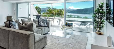 A modern living room with a grey sofa, a glass coffee table, and a view of a lake and mountains through large windows.