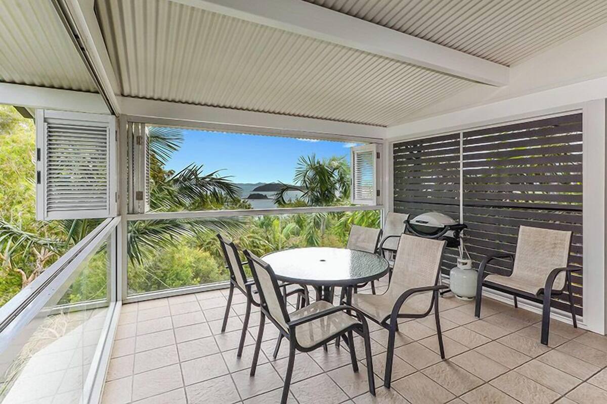 A covered outdoor patio with a round table and chairs, a grill, and a view of greenery and water.