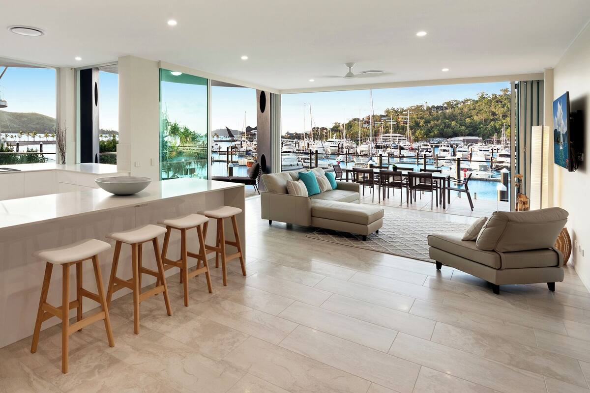 A modern living space with a kitchen island, bar stools, a sofa, and a view of a marina.