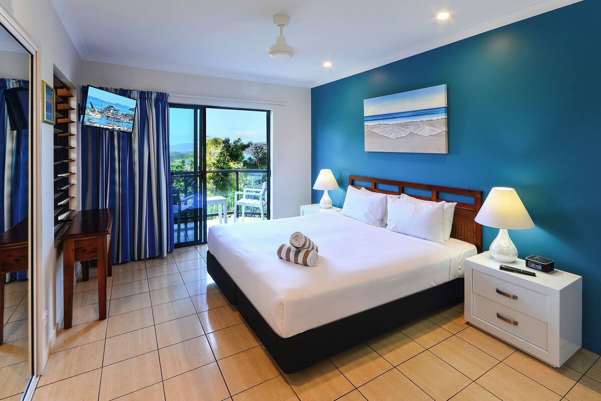 A hotel room with a large bed, two bedside tables with lamps, a view of the sea through the window, a balcony with a chair, and a desk with a chair.