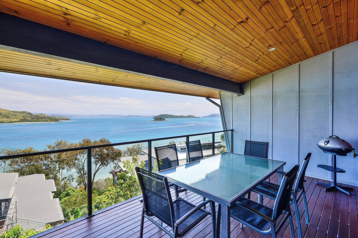 A balcony with a glass table and chairs, overlooking a body of water and distant land.