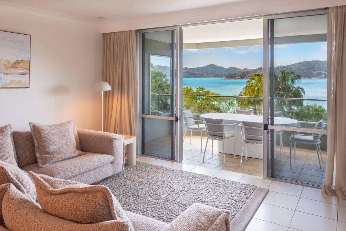 A modern hotel room with a sofa, a dining table, and a view of the sea through large glass doors.