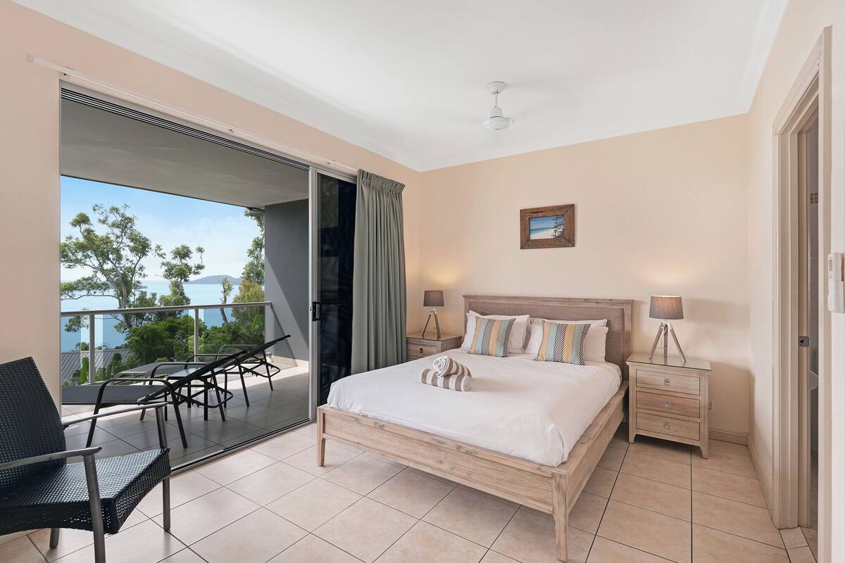 A bedroom with a bed, bedside tables, a chair, a balcony with a view, and a ceiling fan.