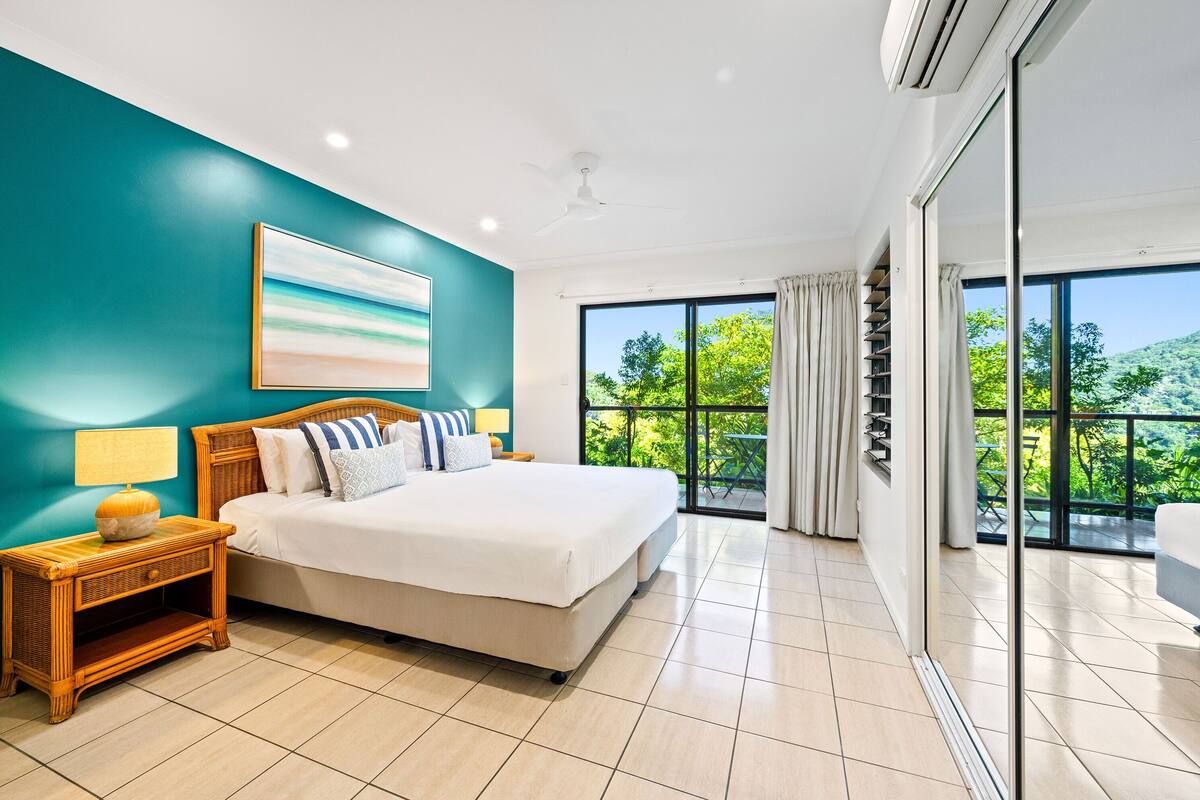 A bedroom with a bed, nightstand, and a large window with a view of greenery.