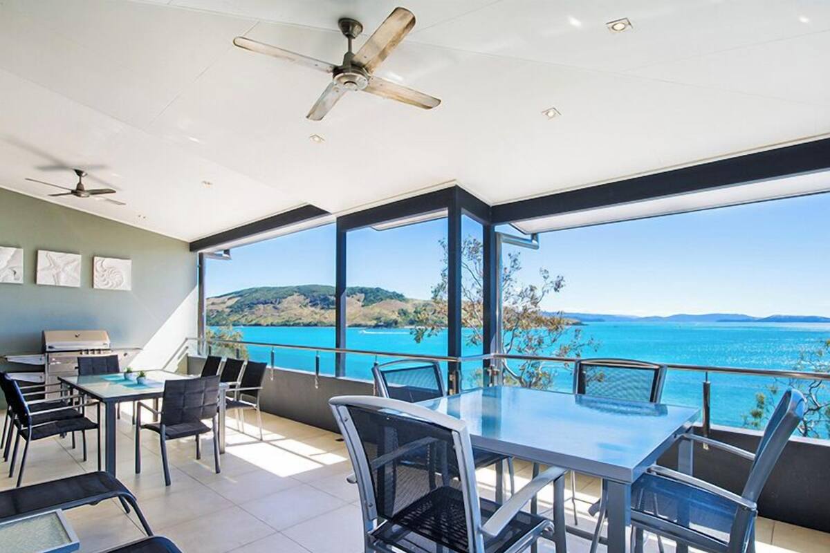 A spacious balcony with a clear view of the sea and hills, a ceiling fan, and a dining area with chairs and a table.