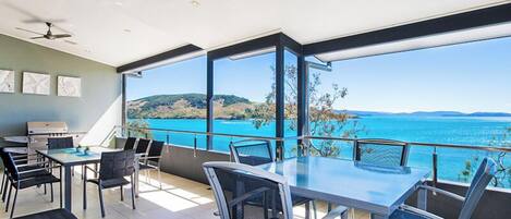 A spacious balcony with a clear view of the sea and hills, a ceiling fan, and a dining area with chairs and a table.