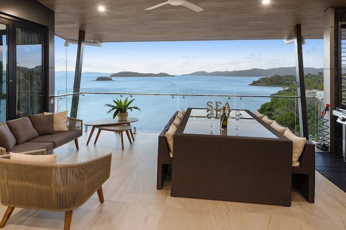 A modern living room with a glass table, a sofa, and a chair, overlooking a body of water and islands.