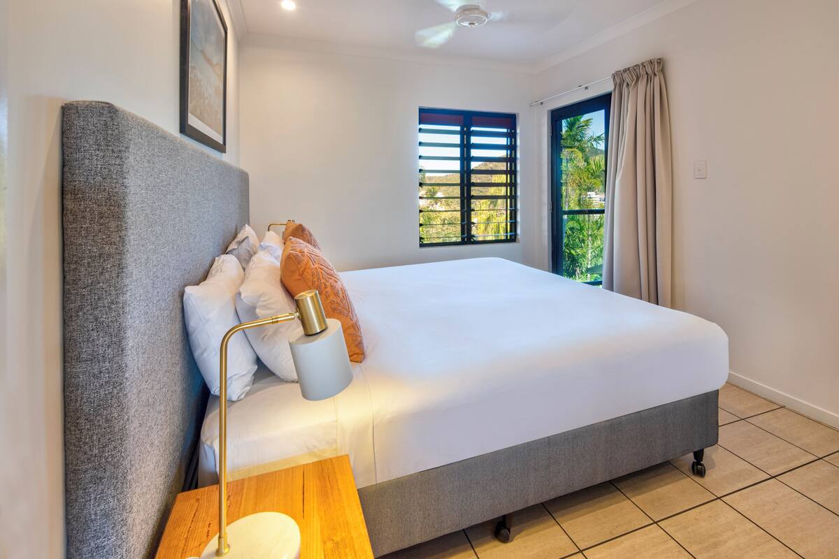 A bedroom with a bed, a bedside table, a lamp, a window with blinds, and a view of greenery outside.