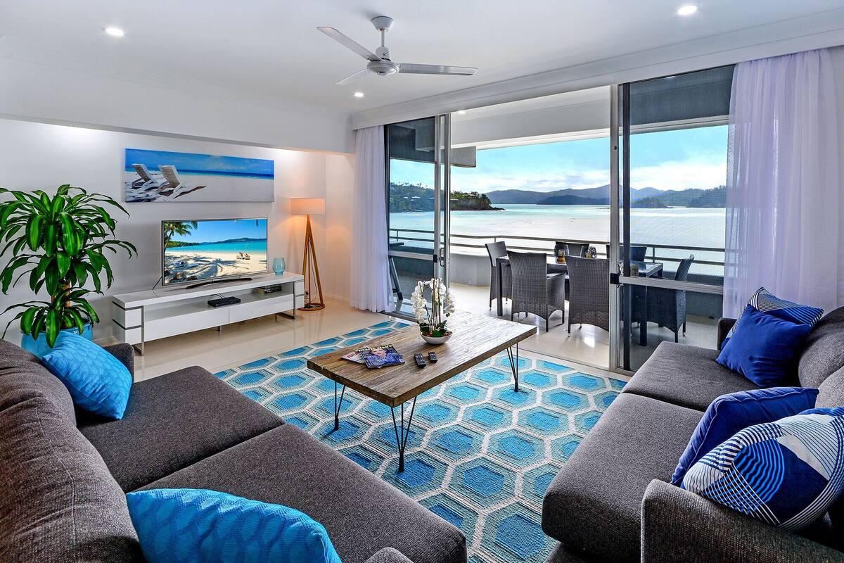 A modern living room with a large TV, a plant, and a balcony with a view of the sea.