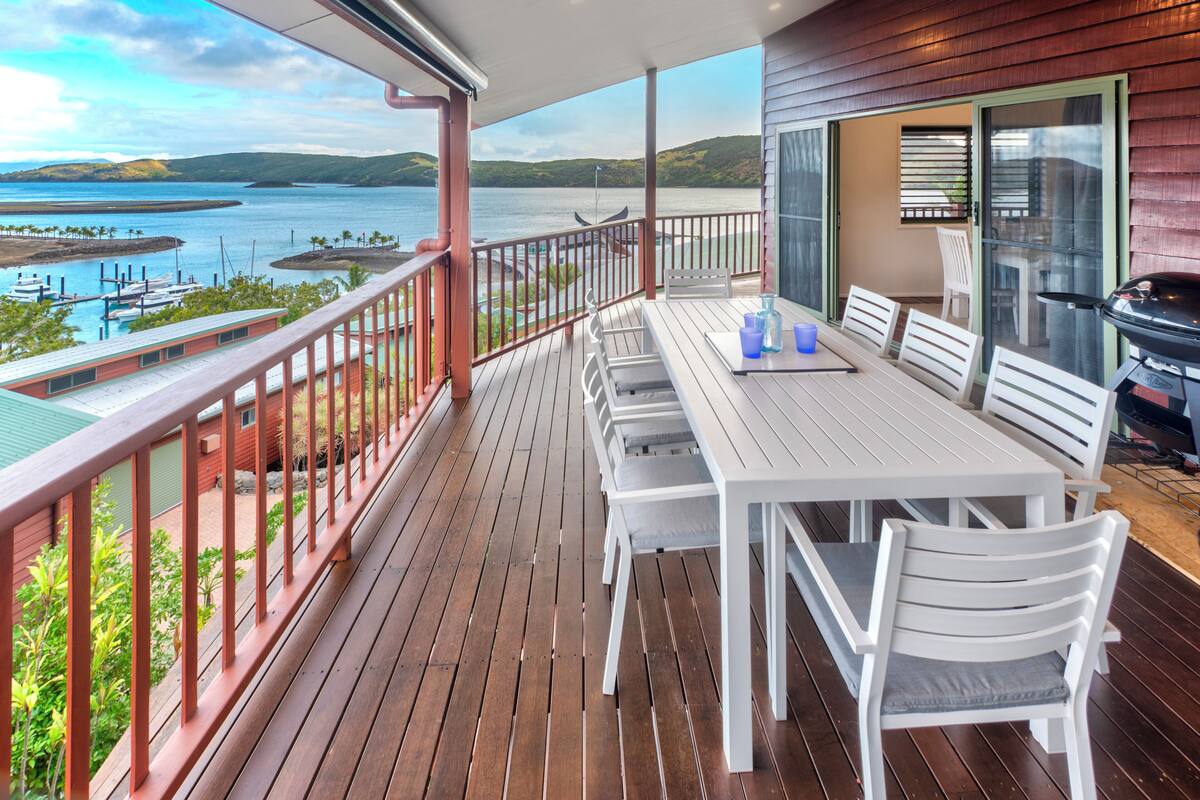 A wooden deck with a table and chairs, a grill, and a view of a lake with boats.